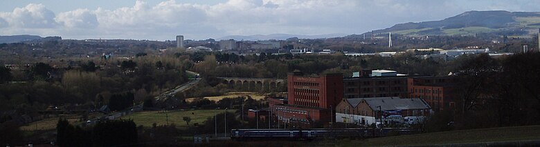 View of Glenrothes seen in its landscape setting from a nearby cemetery. A train is leaving nearby Markinch Station on the East Coast Mainline. Glenrothes town centre with the numerous taller residential and office buildings can be seen in the centre of the image. The River Leven Bridge provides a stark white vertical emphasis on the right side of the image. The Lomond Hills regional park and rolling countryside form the backdrop on the horizon