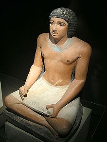 Крашеная, реалистичная, каменная статуя черноволосого мужчины, сидящего со скрещенными ногами и держащего каменное изображение папирусного свитка на коленях