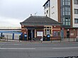 A red-bricked building with a rectangular, dark blue sign reading "KENTON STATION" in white letters all under a blue sky with white clouds
