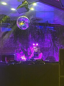 Chelsea Manning, wearing glowing cat ears, workign behind a DJ booth with a discoball overhead