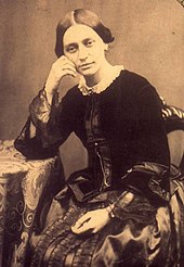 Black-and-white photograph of a woman sitting, looking pensively, with her left hand resting in her lap, while her head leans on her right hand, dressed in a solid-black jacket and lighter skirt