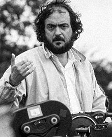 A black and white photograph of a bearded Kubrick dressed in a jacket. He is gesturing with his hand while standing above a large camera.