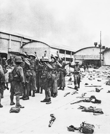 Many British soldiers hold their hands in the air, while Japanese soldiers in the foreground level their weapons at them. Weapons and kitbags litter the ground on the right side of the photo.
