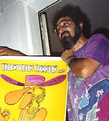 Photograph of a bearded man; he is wearing glasses and a watch, and holds in his hands a colorful poster depicting the comic character Freewheelin' Franklin