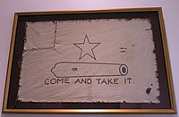 A white flag, with a five-pointed-star over the outline of a small cannon, with the words Come and Take It below. The left corner of the flag has been torn off and sewn back on.