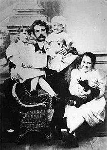 Photograph of a seated man holding two toddlers. At his feet sits a child's rattan rocking chair and a young girl clutching a doll.