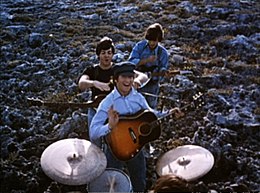 The Beatles performing music in a field. In the foreground, the drums are played by Starr (only the top of his head is visible). Beyond him, the other three stand in a column with their guitars. In the rear, Harrison, head down, strikes a chord. In the front, Lennon smiles and gives a little wave towards camera, holding his pick. Between them, McCartney is jocularly about to choke Lennon.