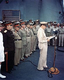 A man in a khaki uniform stands a microphone, reading from papers in his hand. A line of men in smart uniforms stands at attention behind him.
