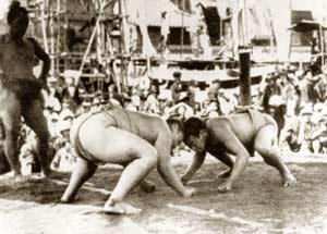 Two sumo wrestlers confront each other on a platform, their heads touching and their fists on the ground. To the side, a third man, also in a wrestling outfit, looks on. In the background, a crowd watches.