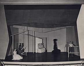 A stage. People sit under the outline of a house on the left; a figure in a black cloak stands atop a stump