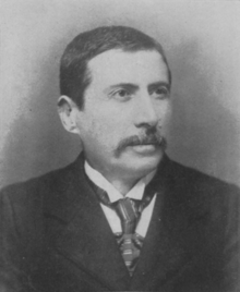 Black-and-white photograph of a white man, in middle age, with short black hair and a moustache. He wears a jacket, collared shirt and tie, and faces slightly to the camera's right.