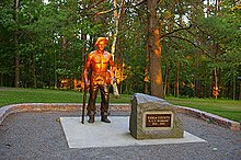 Life-size bronze statue of a shirtless man with a hat, resting his right hand on a pick axe and holding a shirt in his left hand. The top half of the statue is lit orange by the setting sun. A boulder to the right has a plaque that reads "Tioga County 'CCC Worker' 1933 – 1942".