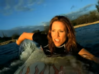 A woman with tan skin swimming on a white surfboard with a visible flame pattern. She is wearing a black wetsuit. Her left hand is submerged in the water. In the background is a beach, and behind it, trees.