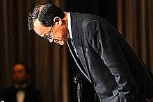 a bespectacled oriental man in a dark suit bowing with brown curtains as backdrop