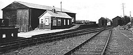 small wooden hut with a large wooden shed behind it. In front of the hut is a platform at two different heights, and in front of the platform are two sets of railway tracks. The tracks lead into a further pair of wooden sheds.