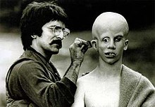 Savini, on the left, applies make-up behind Lehman's ear. Lehman's bald head has been made to appear over-large; his eyes point in directions, and his teeth are extremely crooked.
