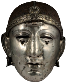 Colour photograph of the Emesa helmet, which has a silver face mask shaped like a human face, and an iron headpiece. The headpiece is decorated with a laurel-wreath diadem and a rosette, and has a central hinge holding the face mask.