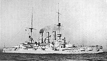 A large warship steaming through calm seas, thin puffs of smoke drift up from her three funnels