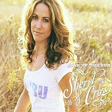A woman with sunlit light brown hair stands in a meadow. She is wearing a white t-shirt with the "BU" of "Malibu" in light blue on the front. The woman faces slightly to the left, with her head turned to the camera. The title at the lower right reads "SOAK UP THE SUN", and below it, "Sheryl Crow" in stylized font.