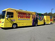 A yellow Winnebago recreational vehicle. Visible on its body is the text "Wheel of Fortune, America's game", as well as photos of Pat Sajak and Vanna White