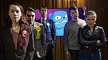Standing in a line: a woman wearing a red badge, a man wearing a yellow badge, a man wearing a blue badge and a blue animated bear on a screen. On the right, an uneasy-looking man stands behind a woman with her arms crossed.