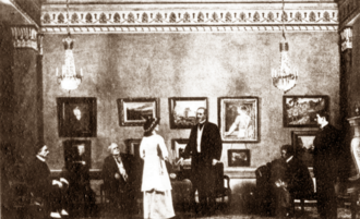 Scene from a play, with young woman standing in a smart drawing room addressing three seated and two standing men