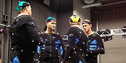 Четири глумца разговарају у оделима за хватање покрета (црна одела која покривају цело тело, заједно са капом на глави, са малим белим лоптицама), у студију